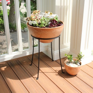 May include: A terracotta planter filled with colorful flowers sits atop a black metal stand with a decorative, twisted design. A smaller terracotta pot with greenery is to the right. The scene is set on a wooden deck.