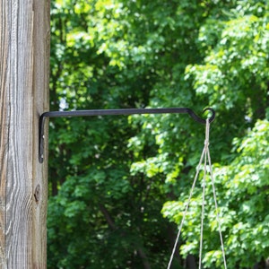 May include: Black metal plant hanger with a hook attached to a wooden post. The hanger is designed to hold a hanging plant pot.