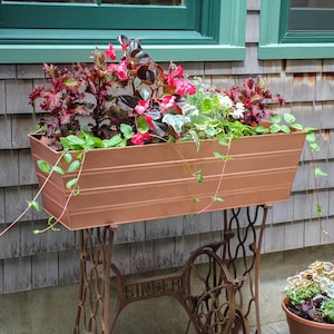 May include: A copper-colored planter box with a variety of pink and green plants sits on top of an antique Singer sewing machine base.