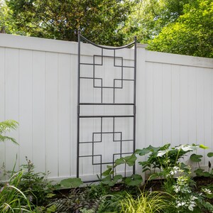 May include: A black metal trellis with a geometric design, standing against a white vinyl fence. The trellis is made of square and rectangular shapes, creating a grid pattern. The trellis is in a garden setting with green plants in the foreground.