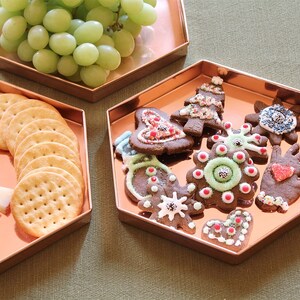 May include: Three copper-coloured hexagonal trays. One holds green grapes, another crackers and cheese, and the third has decorated Christmas biscuits. The biscuits are in various shapes and colours, festive.