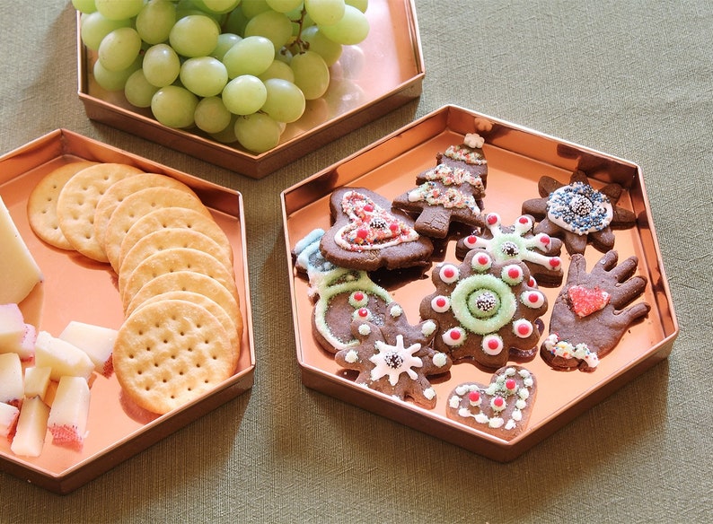 May include: Three copper-coloured hexagonal trays. One holds green grapes, another crackers and cheese, and the third has decorated Christmas biscuits. The biscuits are in various shapes and colours, festive.