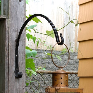 May include: A black metal hook with a twisted design is mounted on a wooden post. A copper lantern hangs from the hook.
