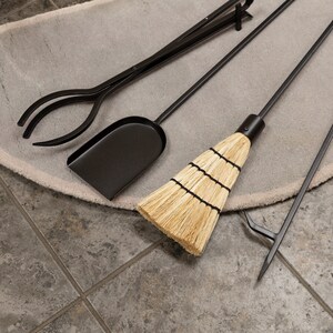 May include: A black fireplace tool set including tongs, a shovel, a broom, and a poker. The tools are arranged on a light gray rug, with a tiled floor in the background. The broom has a natural straw head.