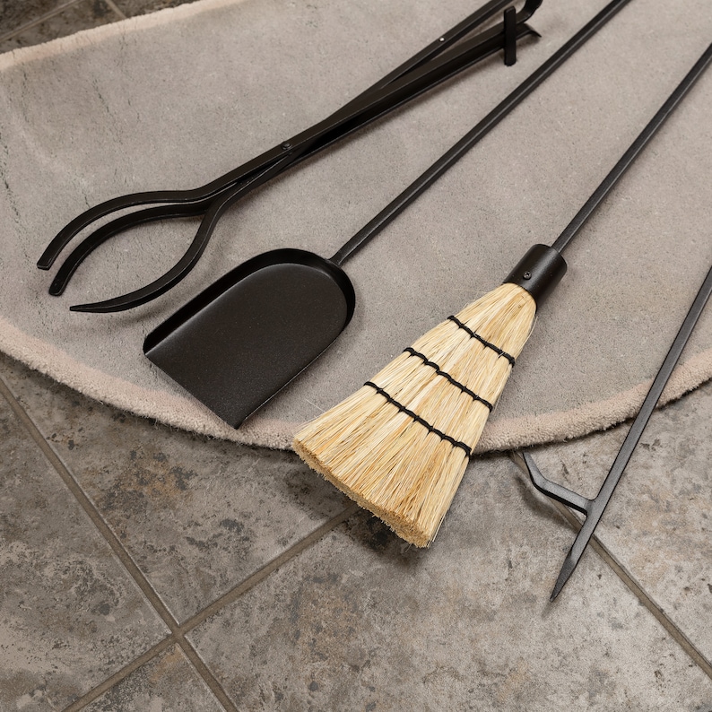 May include: A black fireplace tool set including tongs, a shovel, a broom, and a poker. The tools are arranged on a light gray rug, with a tiled floor in the background. The broom has a natural straw head.