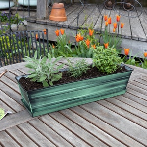 May include: A green metal planter box with a distressed finish, filled with various plants, including sage, rosemary, and oregano. The planter is sitting on a wooden table.