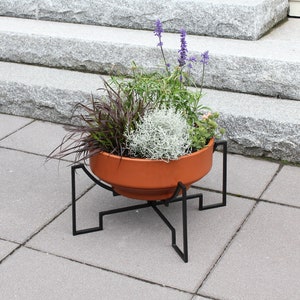 May include: A terracotta planter with a black metal stand. The planter is filled with a variety of plants, including lavender, grass, and a white feathery plant.