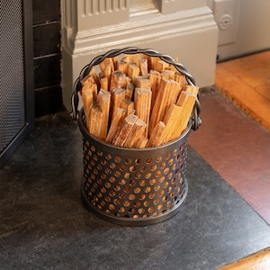 May include: A black metal basket with a wire mesh design holds a pile of wooden kindling sticks. The basket has a black metal handle.