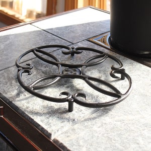 May include: A black metal stovetop grate with a decorative design. The grate is round and has four legs. It is sitting on a gray stone surface.