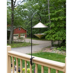 May include: A black metal bird feeder with a clamp for attaching to a railing. The feeder has two brass-coloured bowls, one above the other, and is hanging from a curved metal arm.