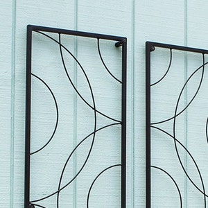May include: Two black metal decorative wall panels with a geometric design of intersecting curved lines. The panels are rectangular and mounted on a light blue textured wall.