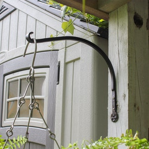 May include: Black metal hanging hook attached to a wooden post. The hook is curved and has a decorative finial. The hook is mounted on a shed with a window.