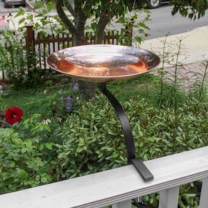 May include: A copper bird bath with a hammered finish sits on a black metal stand mounted to a white wooden railing. The bird bath is positioned in a garden setting with green foliage and flowers.