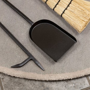 May include: A set of black fireplace tools, including a shovel, poker, and a small broom with straw bristles. The tools are arranged on a gray, circular rug.