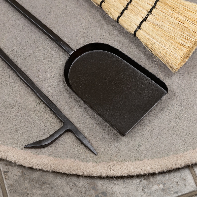 May include: A set of black fireplace tools, including a shovel, poker, and a small broom with straw bristles. The tools are arranged on a gray, circular rug.