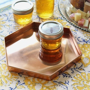 May include: A copper-coloured hexagonal tray holds a small jar of honey. Two additional jars of honey are in the background. The jars have silver lids. The tray sits on a floral patterned tablecloth with yellow and blue flowers.
