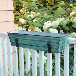 May include: A teal green metal planter box with black metal brackets and hooks. The planter is mounted on a white wooden railing.