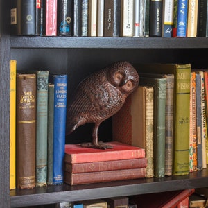 May include: A wooden owl figurine sits on a shelf with a stack of books. The books are mostly brown and have titles like "The Good Earth" and "Through the Looking Glass".
