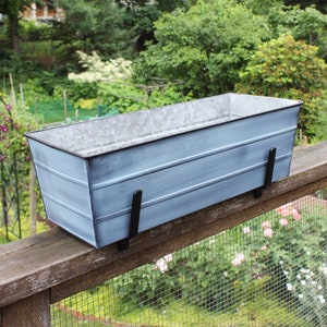 May include: A light blue metal planter box with black metal brackets. The planter is mounted on a wooden railing with a wire mesh fence behind it.