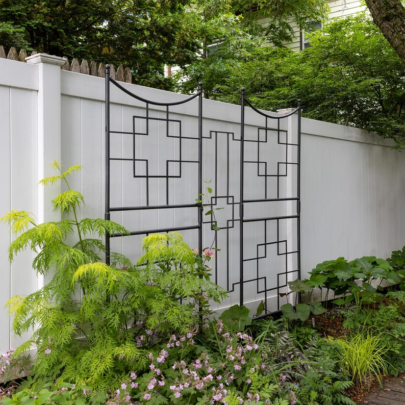 May include: A black metal trellis with a geometric design is attached to a white vinyl fence. The trellis is surrounded by green plants and flowers.