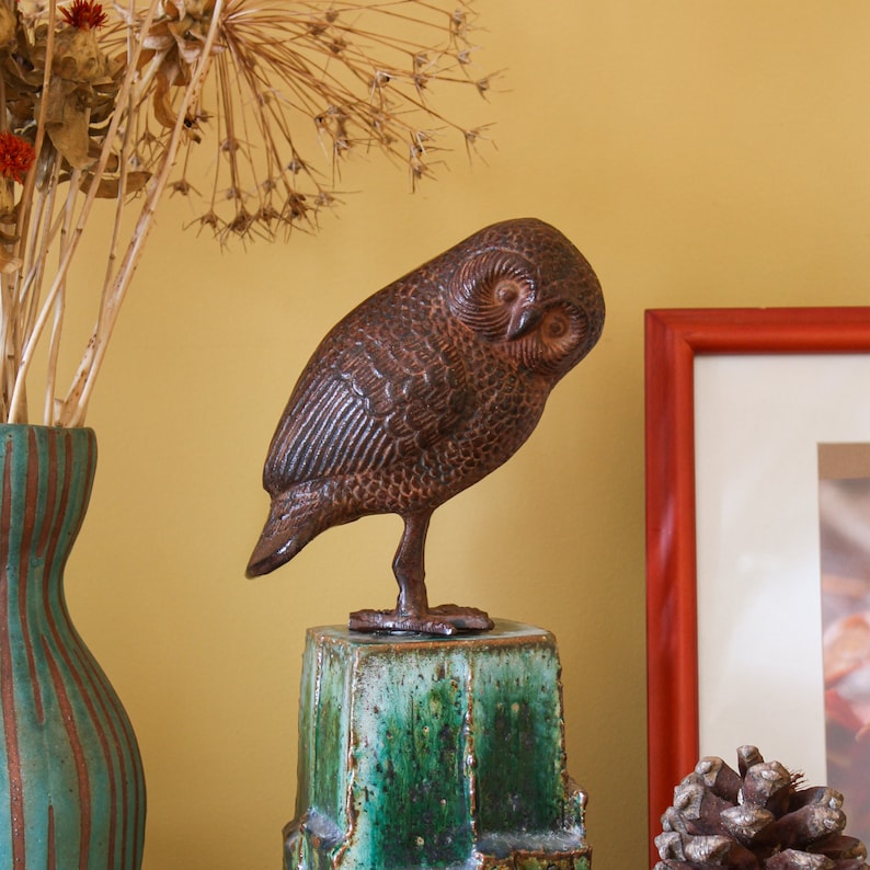 May include: A brown and black owl figurine with a textured surface, perched on a green and blue ceramic base. The owl is positioned on a table with a picture frame and a vase of dried flowers in the background.