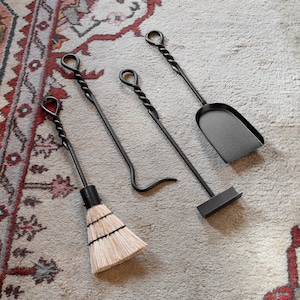 May include: A set of four fireplace tools with black metal handles. The tools include a brush, a poker, a shovel, and a tongs. The tools are arranged on a patterned rug.