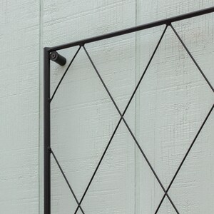 Op de afbeelding: Zwart metalen trellis met een ruitpatroon. De trellis is gemonteerd op een lichtgroene muur.