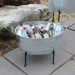 May include: A silver galvanized metal tub with black metal legs filled with various beer and water bottles. The tub is sitting on a gray concrete patio.