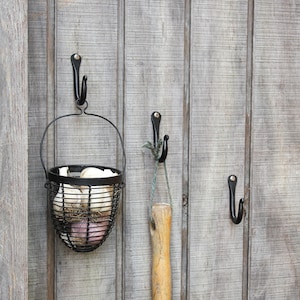 May include: A black metal wire basket filled with garlic bulbs hangs from a black hook on a gray wooden wall. Two other black hooks are also attached to the wall.