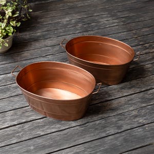 May include: Two oval-shaped copper-colored metal basins with smooth, reflective surfaces and thin wire handles on each side. The basins feature a simple, clean design with horizontal ridges along the exterior walls and a deep, open interior.