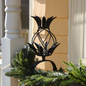 May include: Black metal pineapple-shaped candle holder with a glass votive cup. The decorative piece is placed on a curved stand and surrounded by a green garland. The background features a white column and yellow siding.