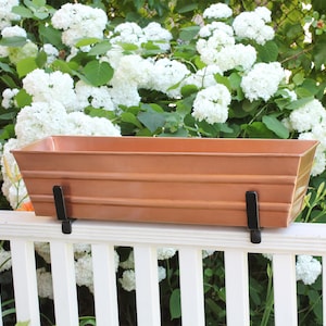 May include: A copper-colored metal planter box with black metal brackets, mounted on a white wooden railing. The planter is empty.