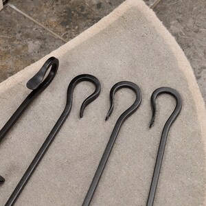 May include: A set of black fireplace tools, including a poker and three curved tools. The tools are made of metal and have a matte finish. The tools are arranged on a light beige rug.