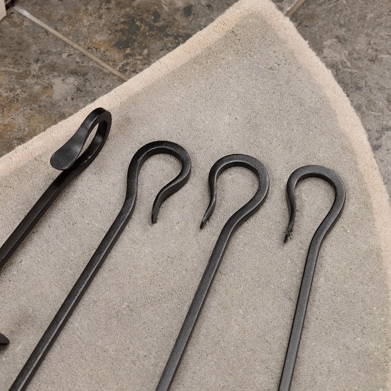 May include: A set of black fireplace tools, including a poker and three curved tools. The tools are made of metal and have a matte finish. The tools are arranged on a light beige rug.