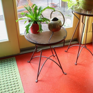 May include: A small, round, metal side table with a perforated top and three curved legs. A potted plant with green leaves sits on the table, next to a small, antique-style watering can. The table is brown and stands on a red floor.