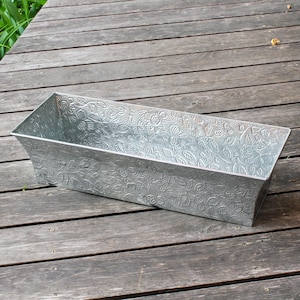 May include: A rectangular galvanized metal planter with embossed floral patterns. The planter has a slightly tapered shape and is designed for outdoor use. The gray metal reflects the sunlight, highlighting the detailed floral design. Perfect for a garden or patio.