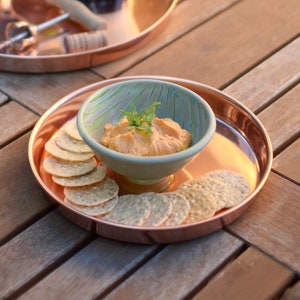May include: A copper serving tray with a small bowl of hummus and a pile of crackers. The bowl is green and white with a leafy design. The tray is on a wooden table.