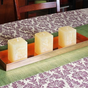 May include: Three ivory-colored candles are arranged in a copper tray on a green and white patterned tablecloth. The candles are square and have a textured surface.