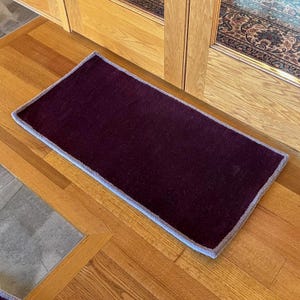 May include: A dark purple doormat with a light gray border. The doormat is lying on a wooden floor.