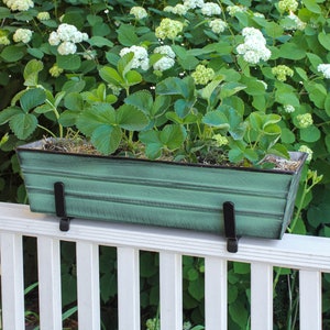 May include: A green metal planter box with black metal brackets, mounted on a white wooden railing. The planter is filled with green leafy plants.