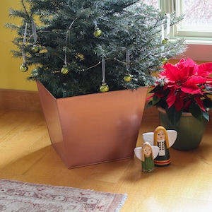 May include: A potted Christmas tree with green ornaments and a copper-colored pot. A red poinsettia plant is in a brown pot next to the tree. Two wooden angel figurines are on the floor in front of the plants.