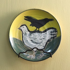 May include: A yellow ceramic plate with a hand-painted design of a black crow perched on a white chicken. The plate has a green and blue border and the artist's signature "Buchita" is visible in the lower right corner.