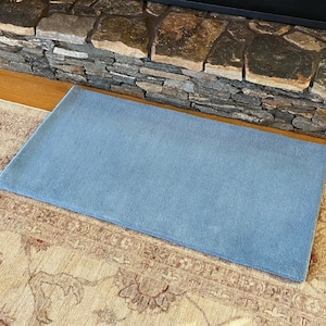 May include: A light blue rectangular rug with a soft, plush texture. The rug is placed on top of a patterned rug with beige and brown tones.