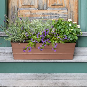 May include: A copper-coloured metal planter box filled with a variety of flowers in bloom, including lavender, white flowers, and purple flowers. The planter is sitting on a wooden deck with a green painted railing.