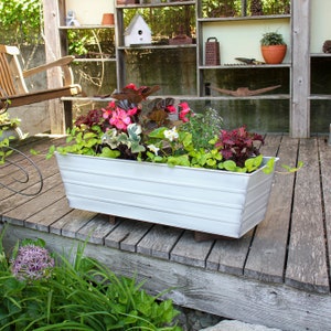 May include: A white metal planter box with a variety of flowers and plants, including pink begonias, green ivy, and white flowers. The planter is sitting on a wooden deck.