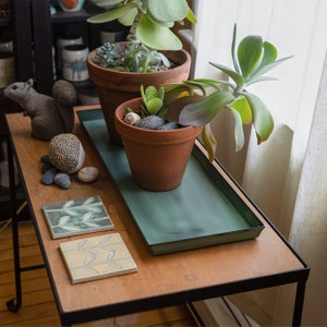May include: A wooden table with a black metal frame, featuring two potted succulents. One pot is brown and the other is green. The table also has two decorative tiles with a leaf design.
