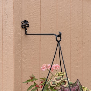 May include: Black metal plant hanger with a triangular design. The hanger is attached to a wall with a decorative hook. A potted plant with pink and white flowers is hanging from the hanger.