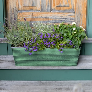 May include: A green metal planter box filled with a variety of flowers, including lavender, white flowers, and purple flowers.
