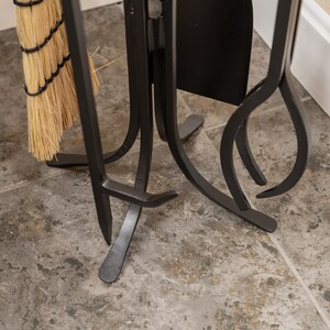 May include: A black fireplace tool set with a matching stand. The set includes a poker, shovel, and brush. The stand has a decorative, curved design. A natural-colored broom is attached to the stand.