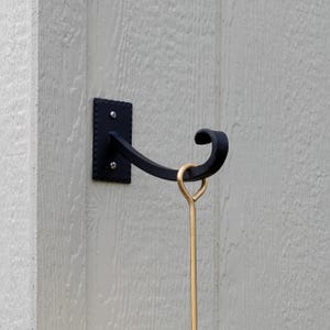 May include: Black wrought iron wall hook with a decorative scroll design. The hook is mounted on a rectangular plate with rounded edges and is secured with two screws. A gold-colored metal rod hangs from the hook.